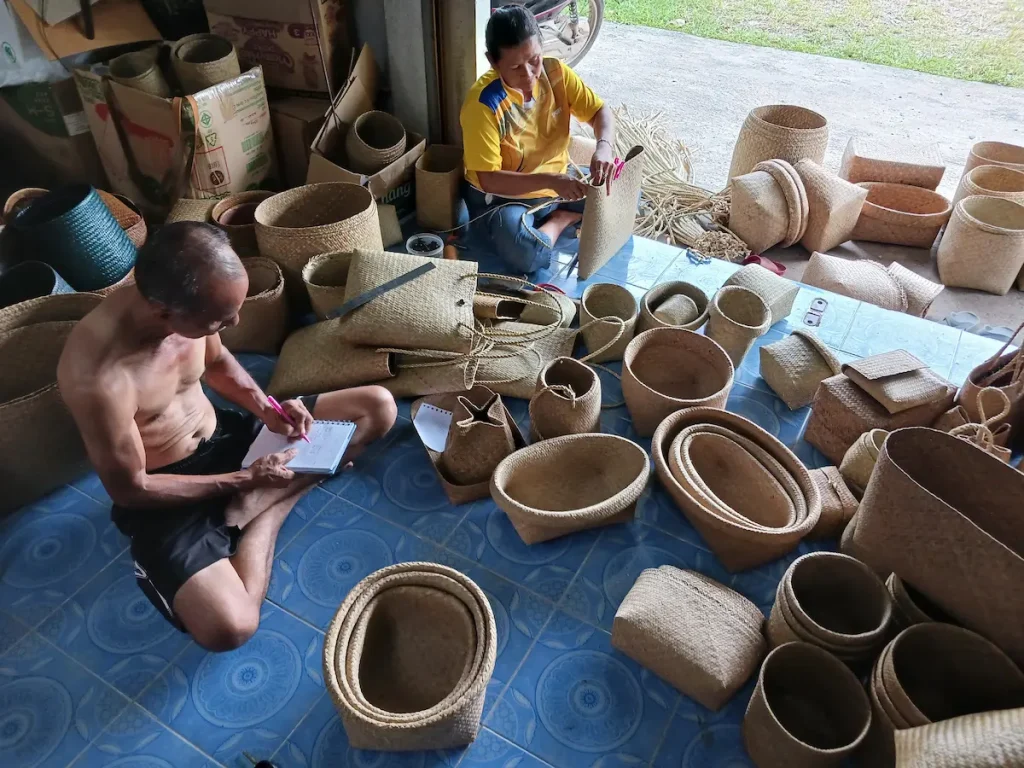 カチュー(水草の一種)のかばんやバスケットに囲まれたアトリエで伝票を整理する夫と注文に合わせてかばんに紐を結び付けている妻
