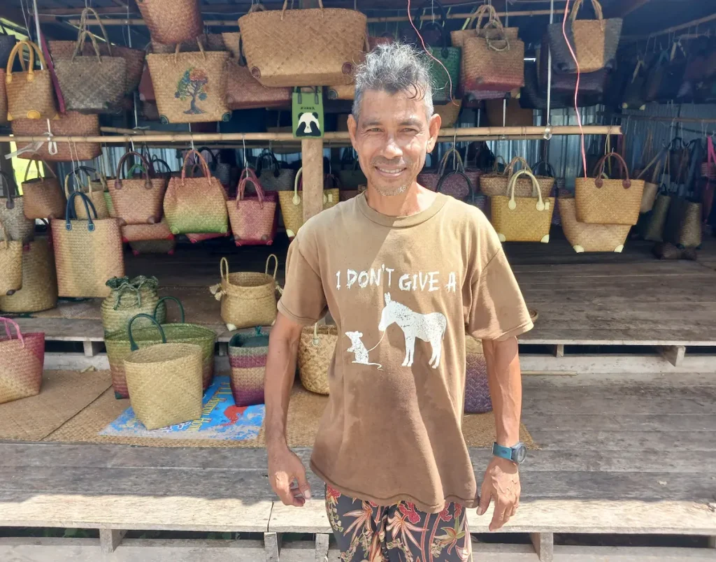 タレノーイ湖周辺の作業場で、様々な色や形のカチュー(水草の一種)編みのカバンを背景にカメラに向かって穏やかに微笑む水草の職人