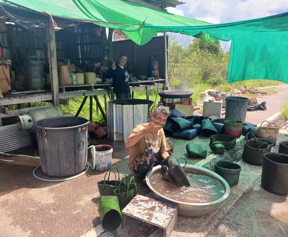 染色したカチュー（水草の一種）のカバンを大きなタライで水洗いする職人夫婦。カメラに向かって笑顔でピースサインをしている