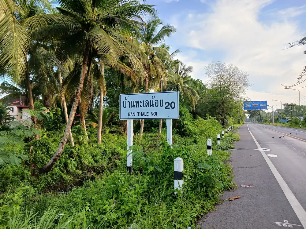 白地にBAN THALE NOI　20と書かれた標識。英語の下にタイ語も表記。
バックに広がる緑と青空
