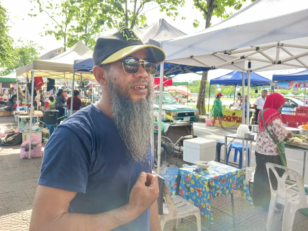 ベースボールキャップにサングラス姿で、顔の倍ほどある顎ひげを触りながら屋台に立つ男性