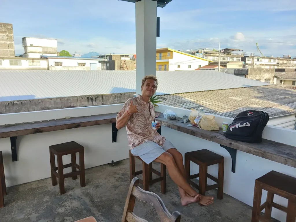 朝日が差し込むゲストハウスの屋上テラスで、朝食を楽しみながらカメラに笑顔を向ける外国人青年