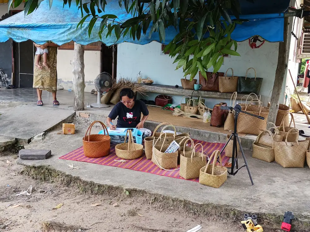 軒先に並べられた様々な色、形のカチュー(水草の一種)のカバンの傍らであぐらをかいて座り、事務作業をしている男性