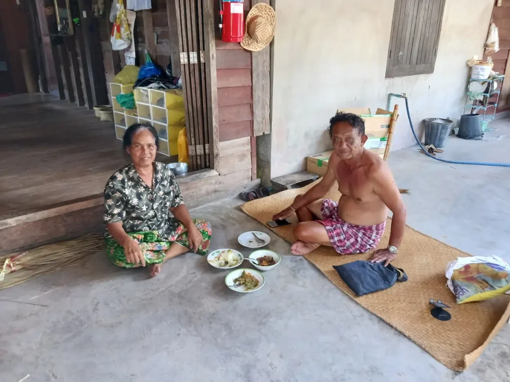 床にあぐらをかいて昼食をとる水草職人の夫婦。皿が床に並び、夫は上半身裸、妻は柄シャツ