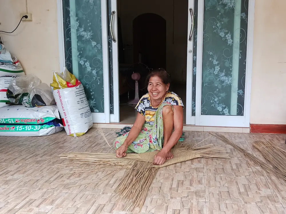 水草の村の軒先でカチュー（水草）を編む女性。片膝を立ててカチューを押さえながら丁寧かつ素早く編み込んでいる
