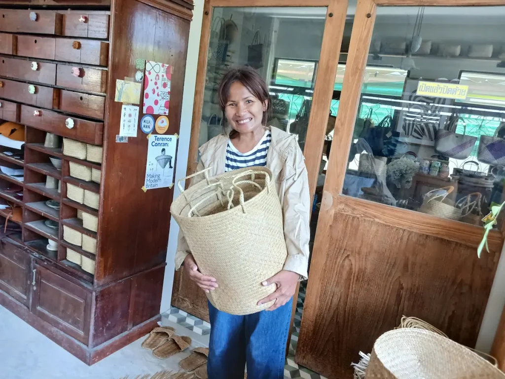 アトリエで背丈の半分ほどもあるカチュー(水草の一)の持ち手付き収納バスケットを両手で持ち上げるカメラに微笑むタイの女性