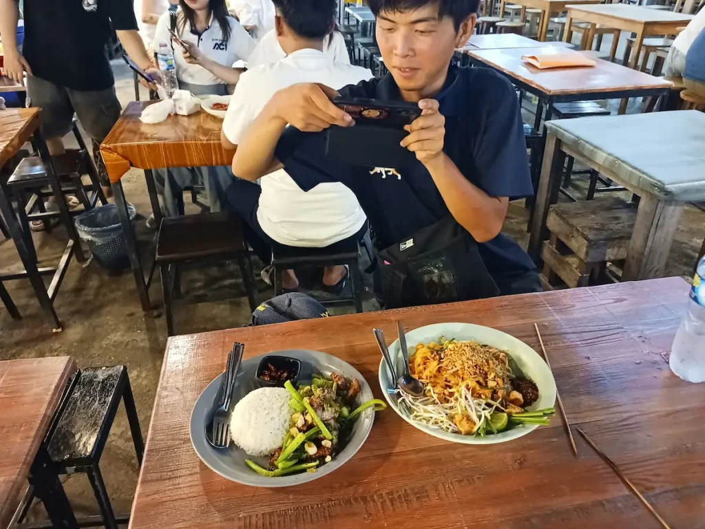 ビエンチャンのナイトマーケットで机の上に置かれた料理2皿とそれを撮影する半袖の男性